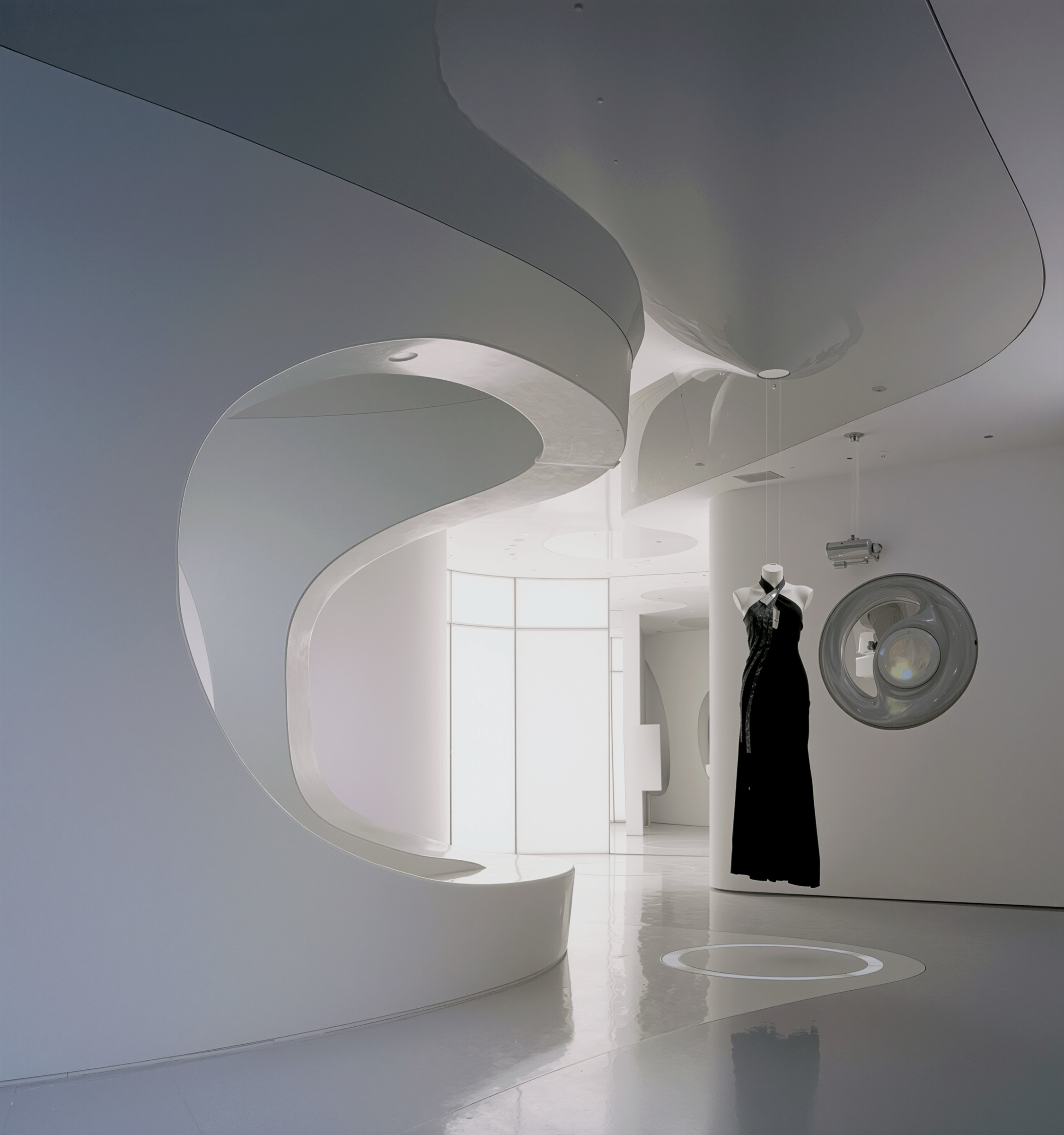 Interior with sweeping white curvilinear ceiling and wall form descending to the floor, a black evening dress on a mannequin, and a circular glass light fixture