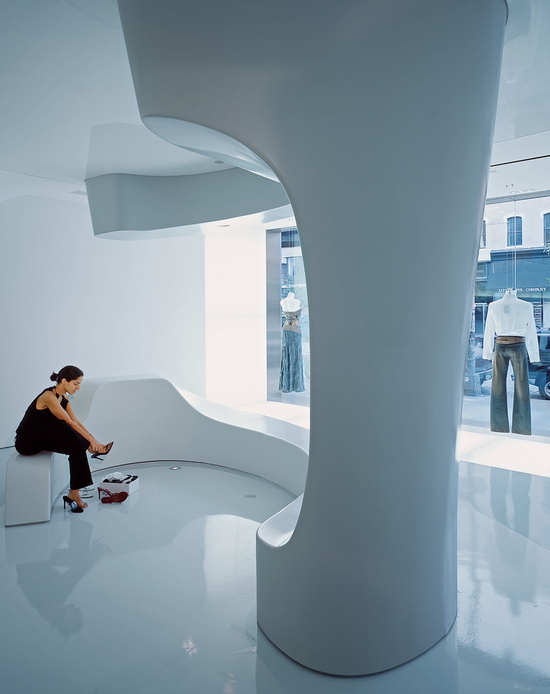Woman seated on a sculptural white bench beside a sweeping column, trying on shoes, with storefront glazing and garments on display visible beyond