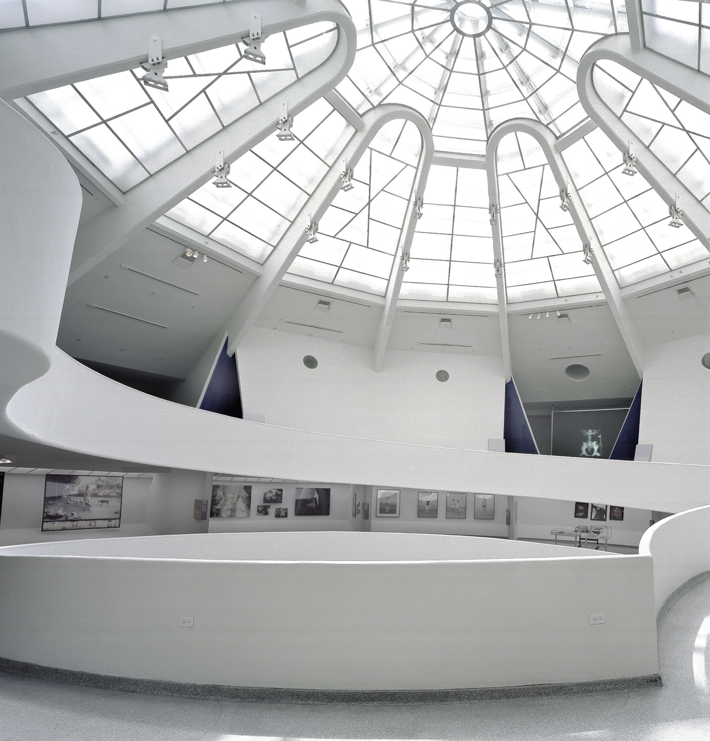 View up through the Guggenheim rotunda toward the skylight — ramps with photographs and blue acoustic panels visible on the upper levels