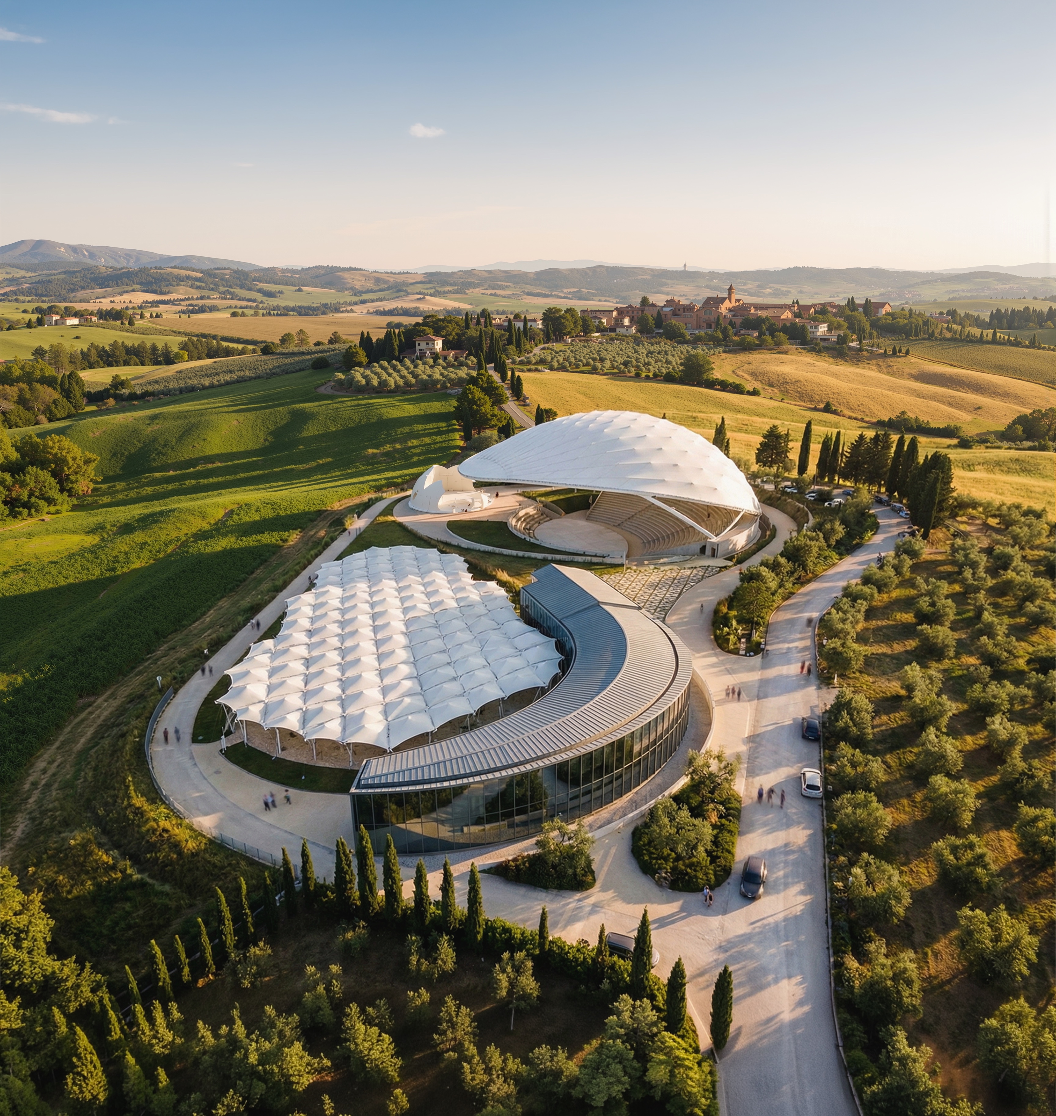 Aerial daytime view of the complete park complex showing the interconnected white-roofed pavilions surrounded by green Tuscan countryside and the town of Peccioli beyond