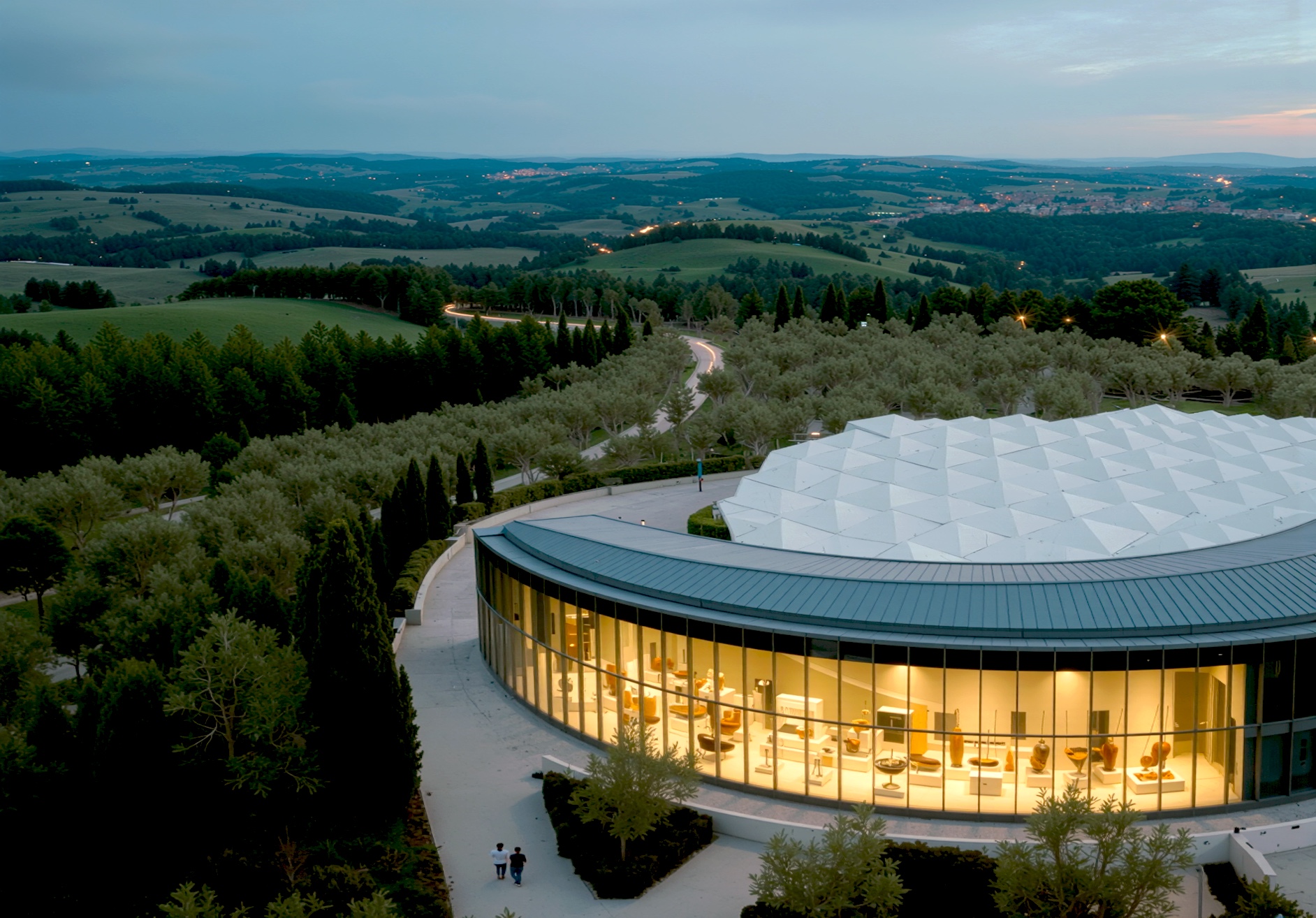 Twilight view of the curved building exterior with its faceted white roof and warmly lit glass ground floor revealing the interior spaces