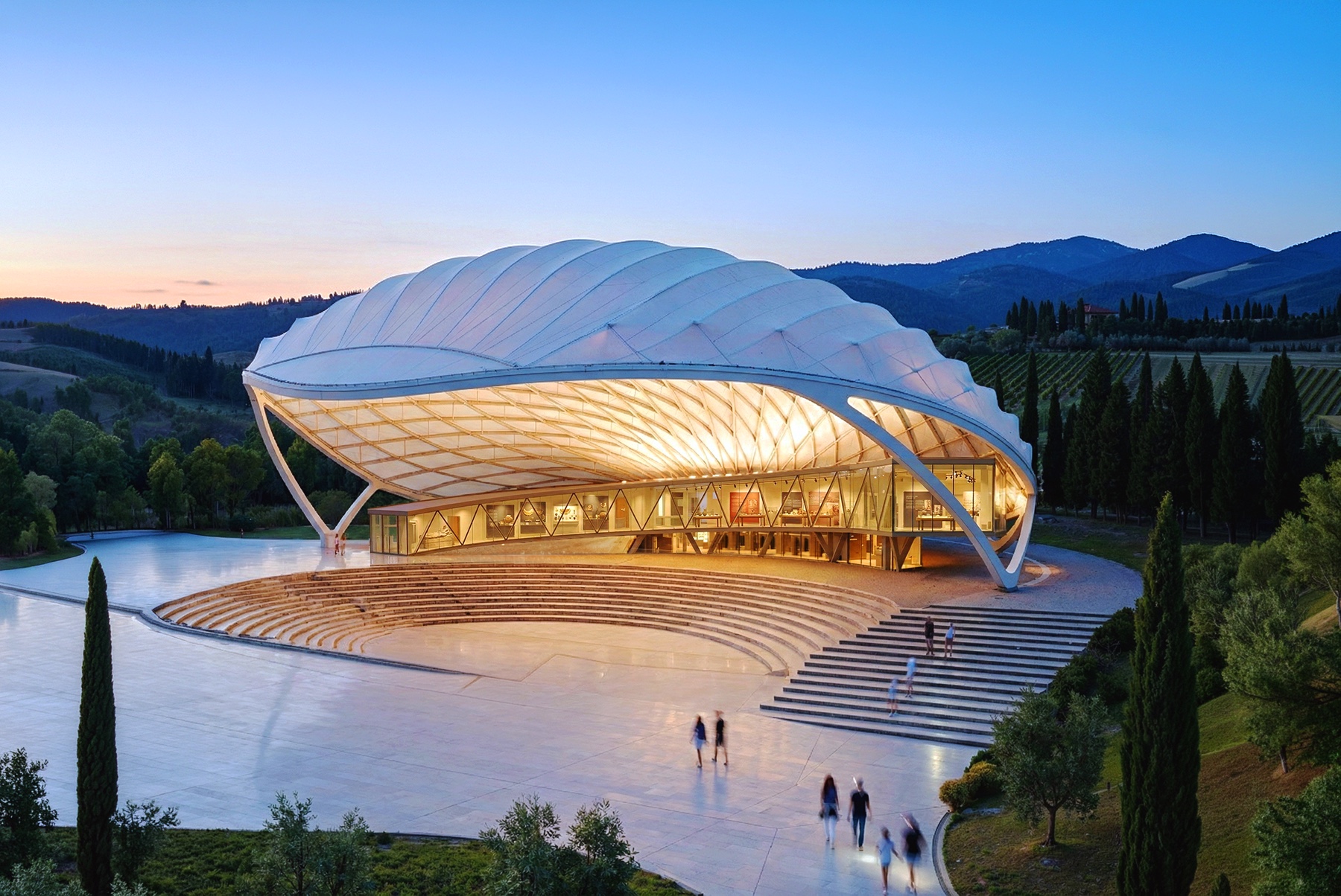 Wing-shaped pavilion structure glowing warmly at dusk with its sweeping white canopy roof and stepped amphitheater seating