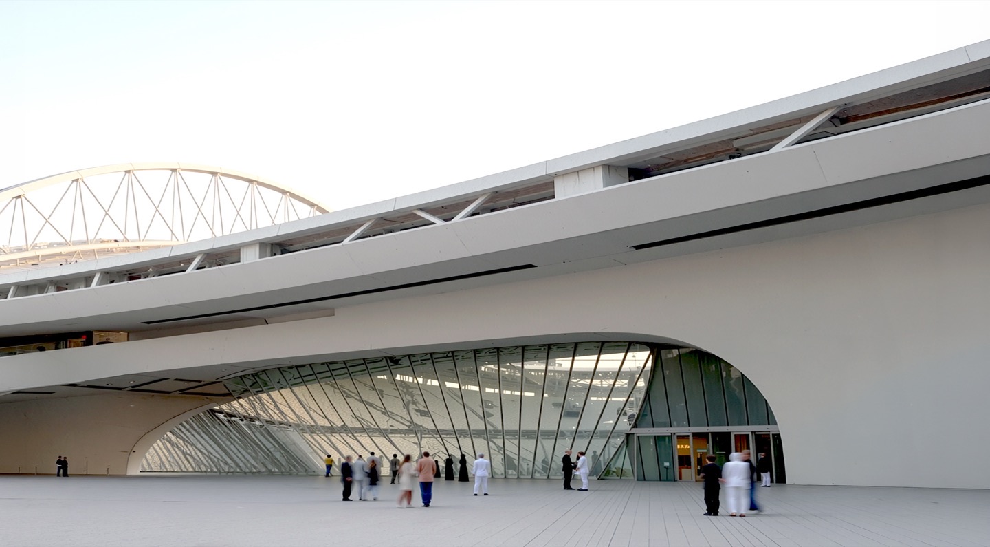Ground-level view of the arena entrance concourse with sweeping curved white roof overhangs, glazed facades, and visitors gathering beneath the structure