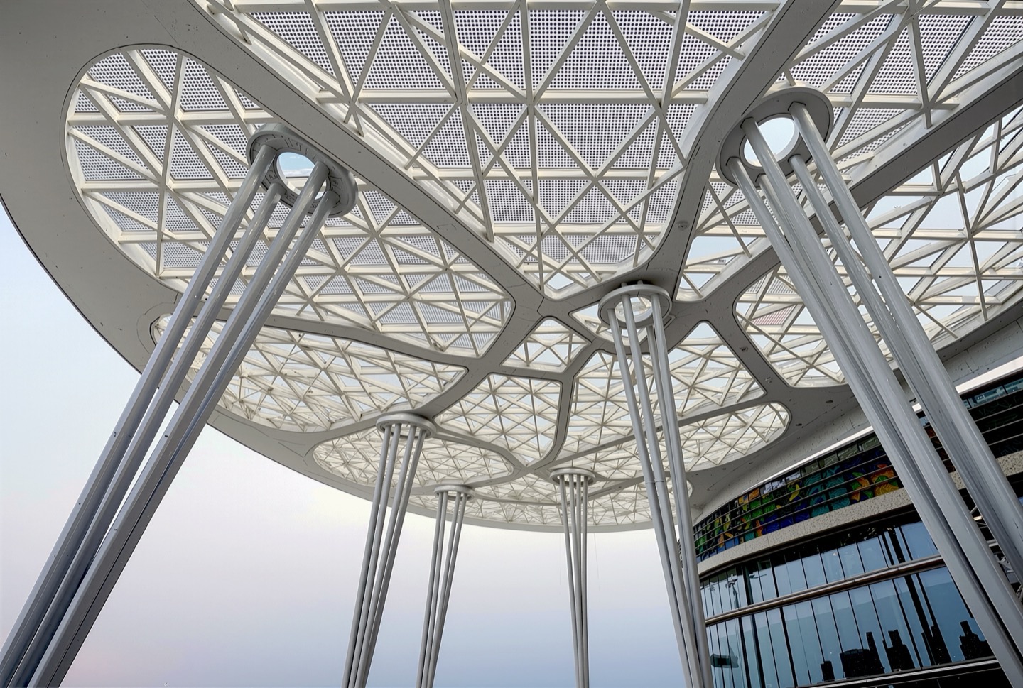 Upward view of the grand entry canopy showing an intricate triangulated steel lattice structure supported by tree-like branching columns against a twilight sky
