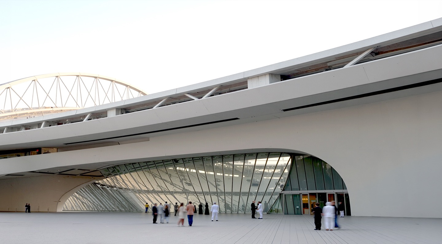 Ground-level view of the arena entrance concourse with sweeping curved white roof overhangs, glazed facades, and visitors gathering beneath the structure
