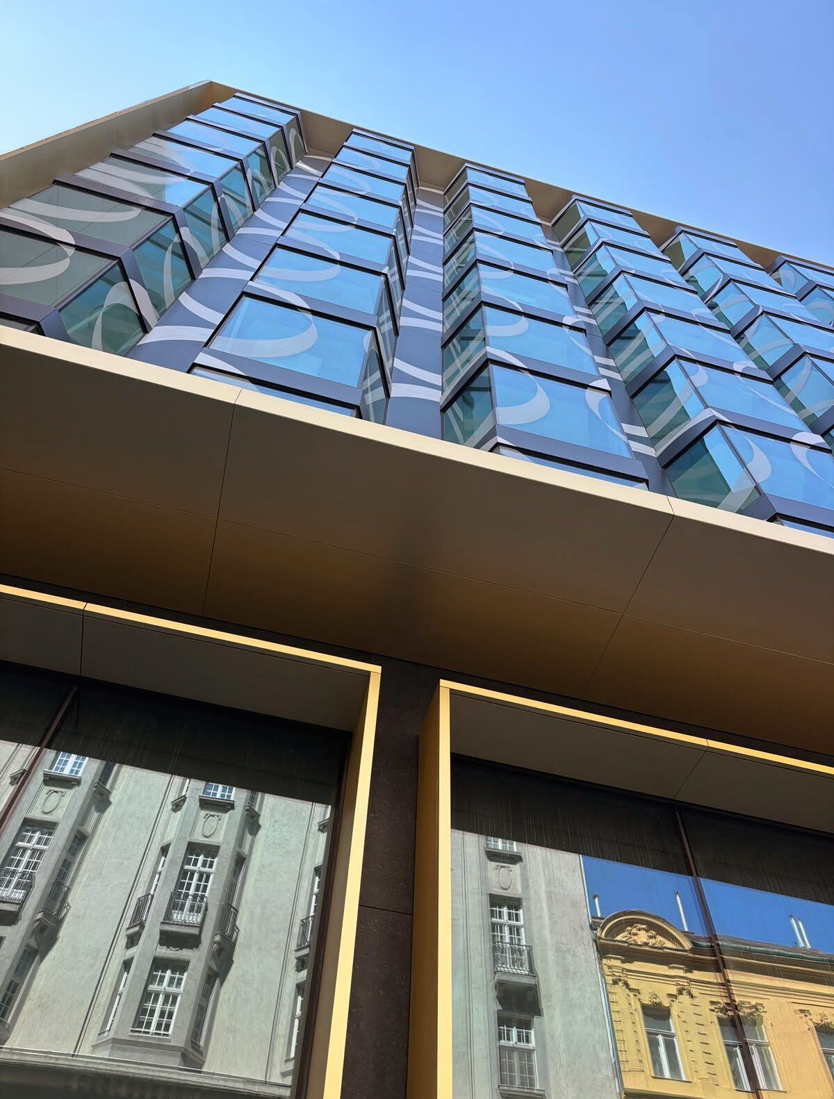 Looking up at the completed SO/ Budapest facade, Op Art circles printed across the angled bay-window glass against clear blue sky, with the bronze fascia and reflected historic Pest neighbours below