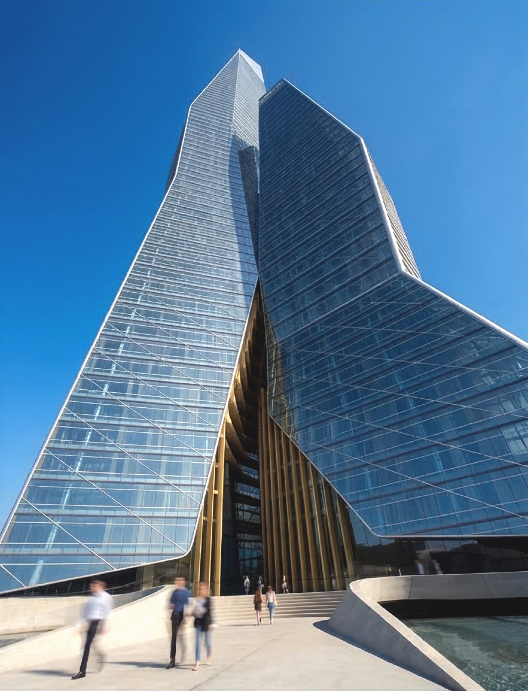 Ground-level view at the base of Solomon Tower, the twin faceted glass volumes rising from a central entry colonnade with people crossing the plaza