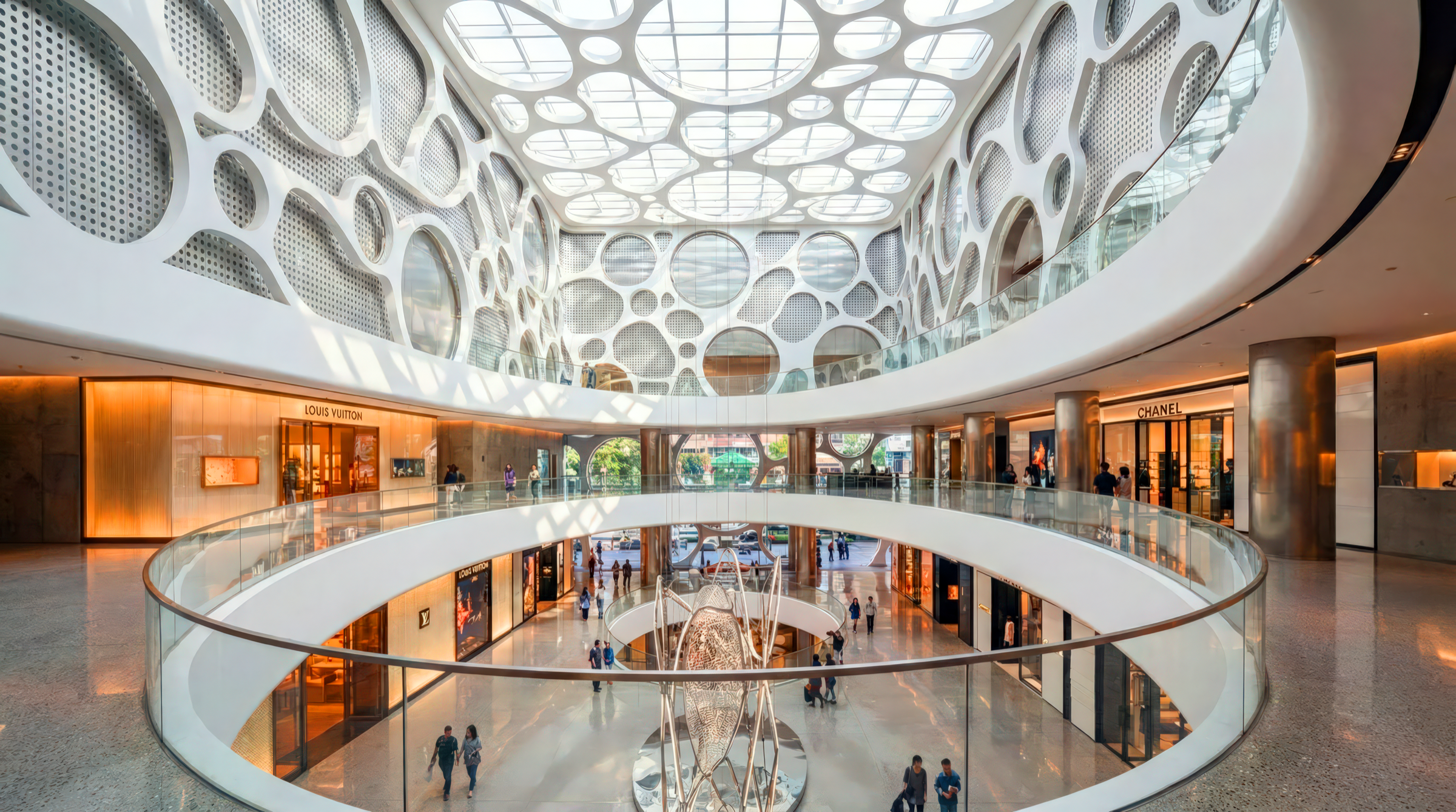 Upper-level interior view of the atrium with the organic cellular dome overhead, a sweeping elliptical glass balustrade, polished stone floors, and luxury storefronts including Chanel visible on the level below