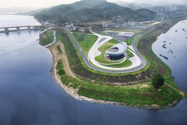 Aerial view of The ARC on its river peninsula showing the circular vessel form, surrounding landscaped pathways, the Nakdong River on both sides, and mountains in the distance
