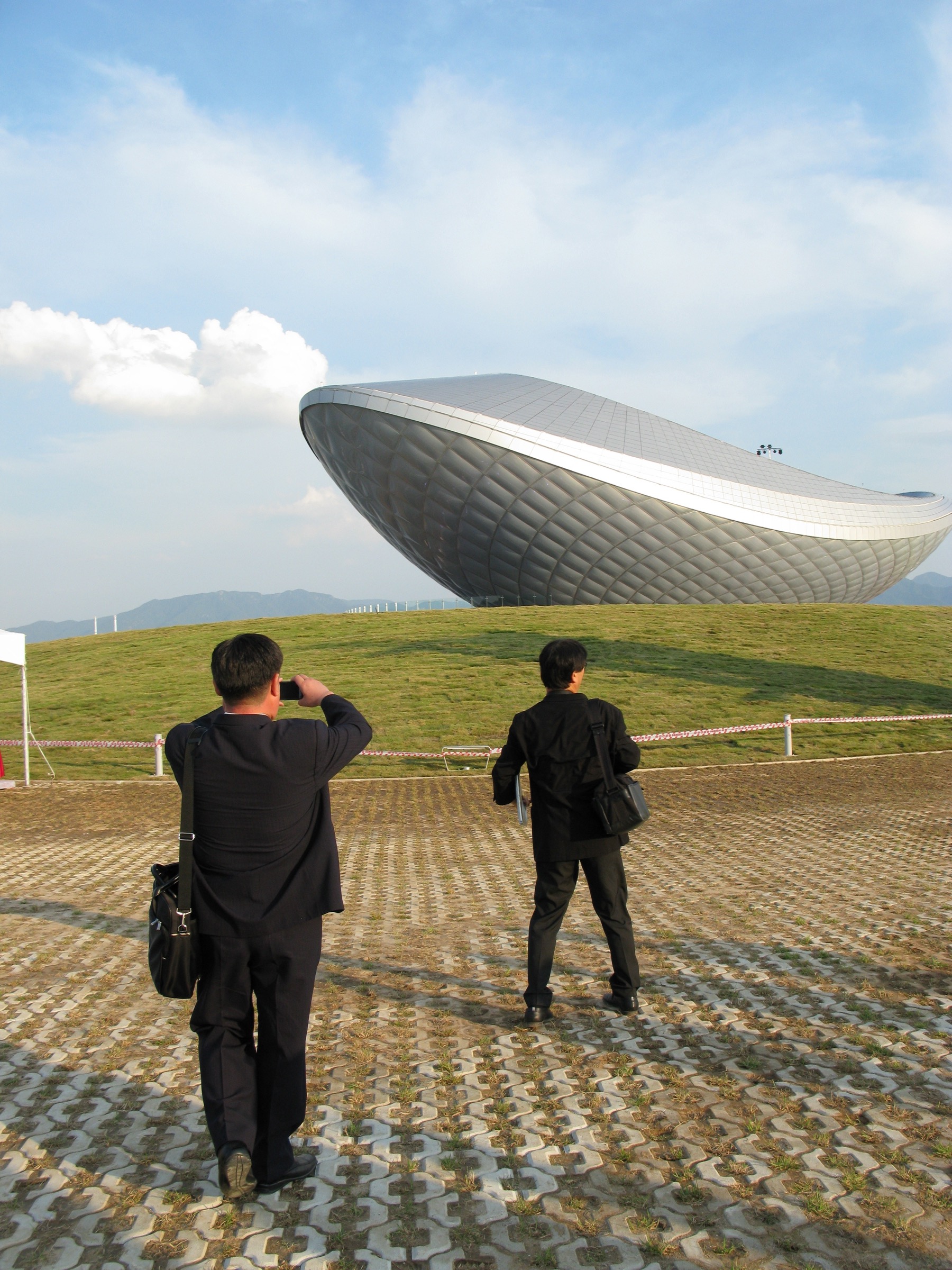 Two visitors photographing The ARC from the approach plaza, the silver vessel form rising from the green landscaped berm against a cloudy sky