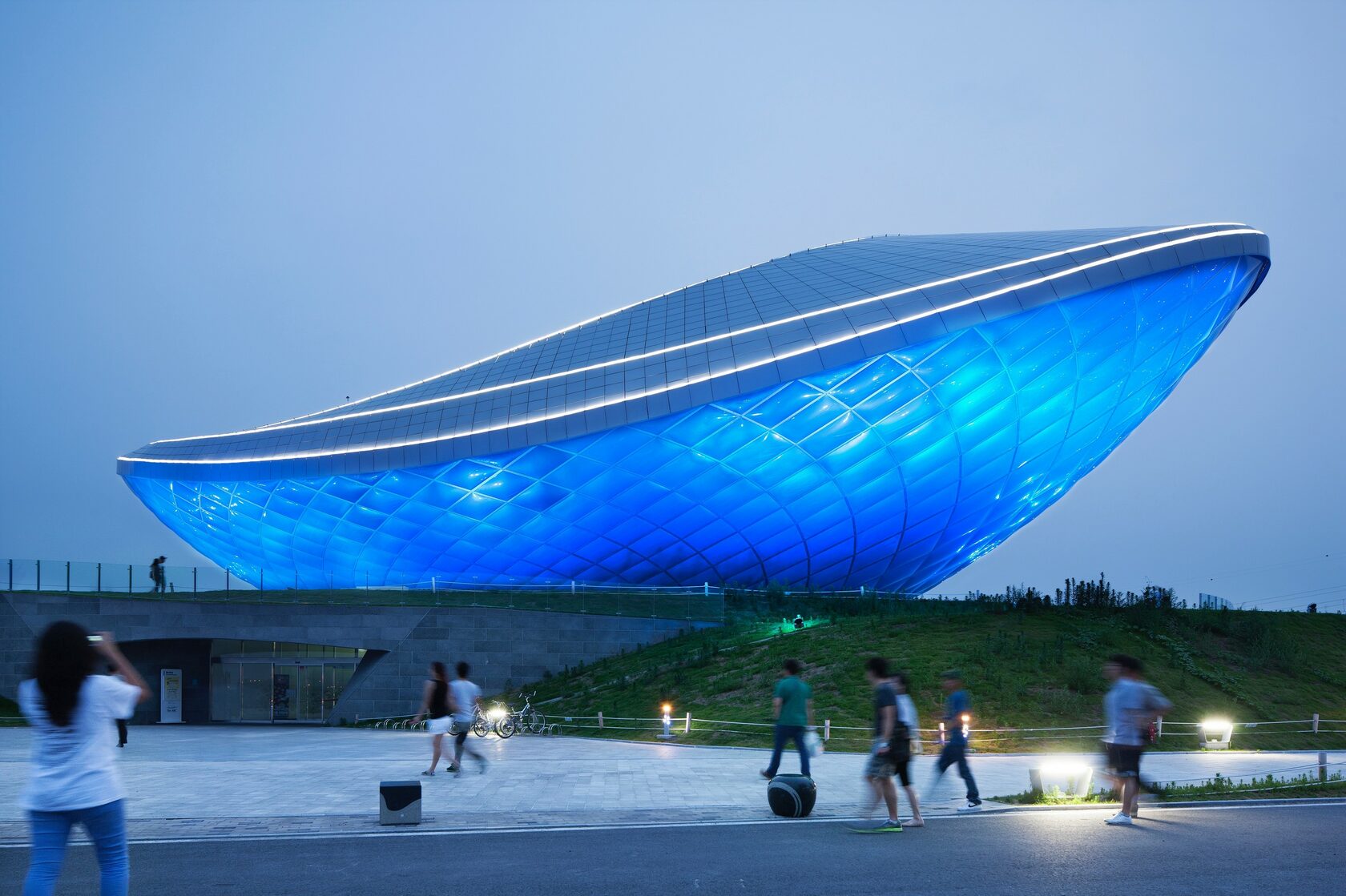 The ARC at dusk with its ETFE-clad vessel form illuminated in blue light, visitors in motion on the plaza, and the landscaped berm rising to the building's edge