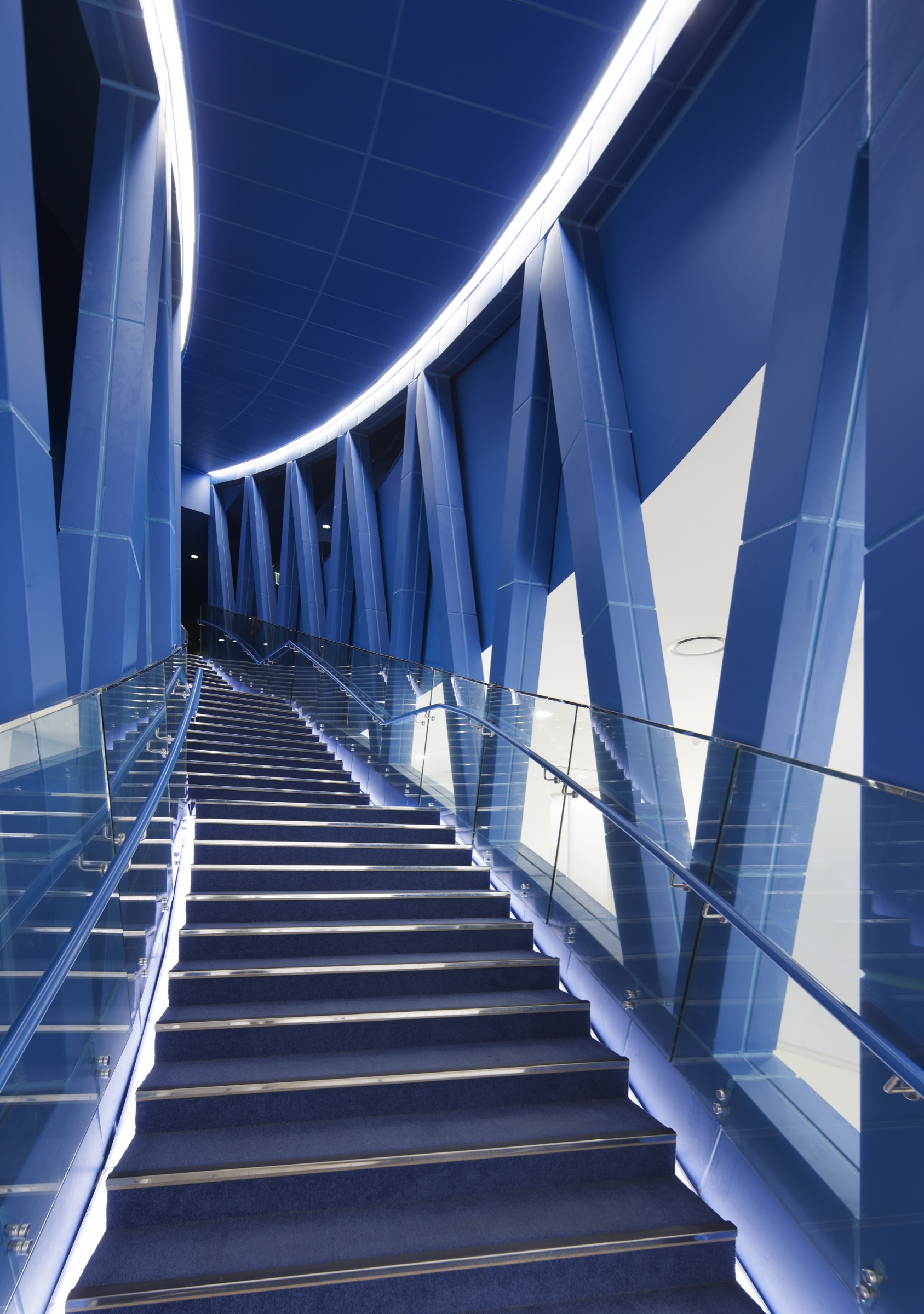 Interior blue-lit staircase ascending between angular blue-clad walls with a curving LED strip tracing the ceiling edge and glass balustrades