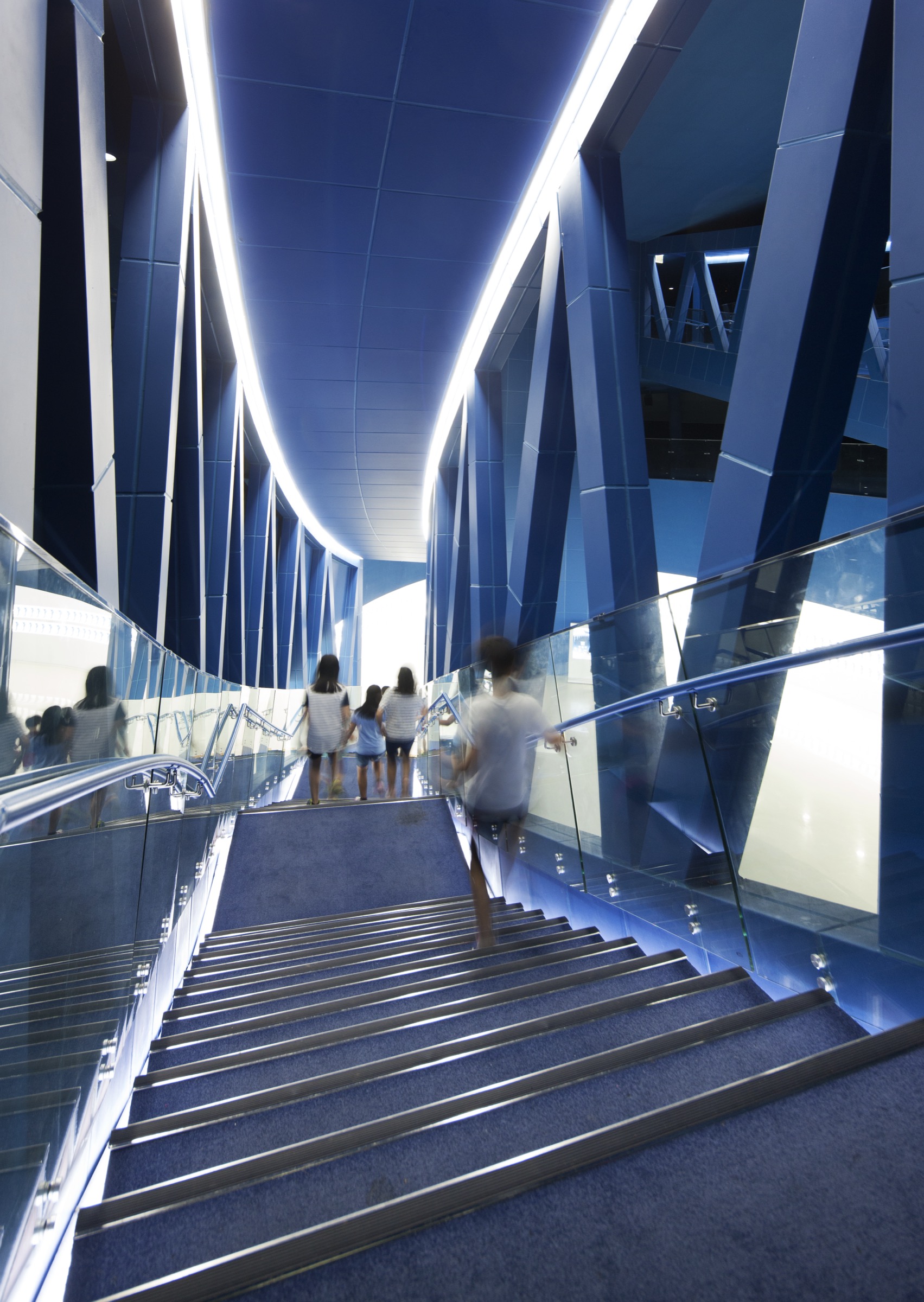 Visitors ascending the blue-lit staircase with daylight visible at the top, angular wall surfaces, and glass handrails