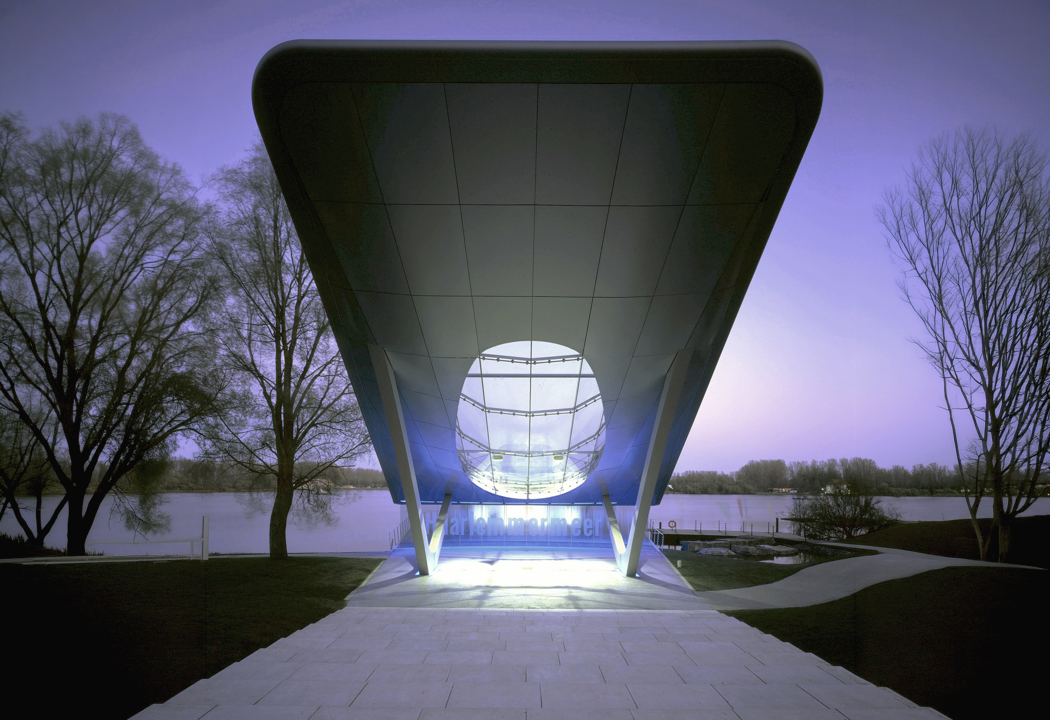 The HydraPier Pavilion at twilight with its dramatic cantilevered roof framing a circular glass aperture, set against the Haarlemmermeer lake