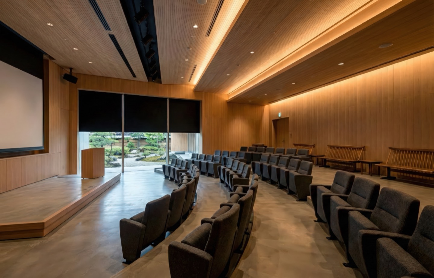 Wood-paneled auditorium with warm timber ceiling, tiered seating, and a garden view through the stage window