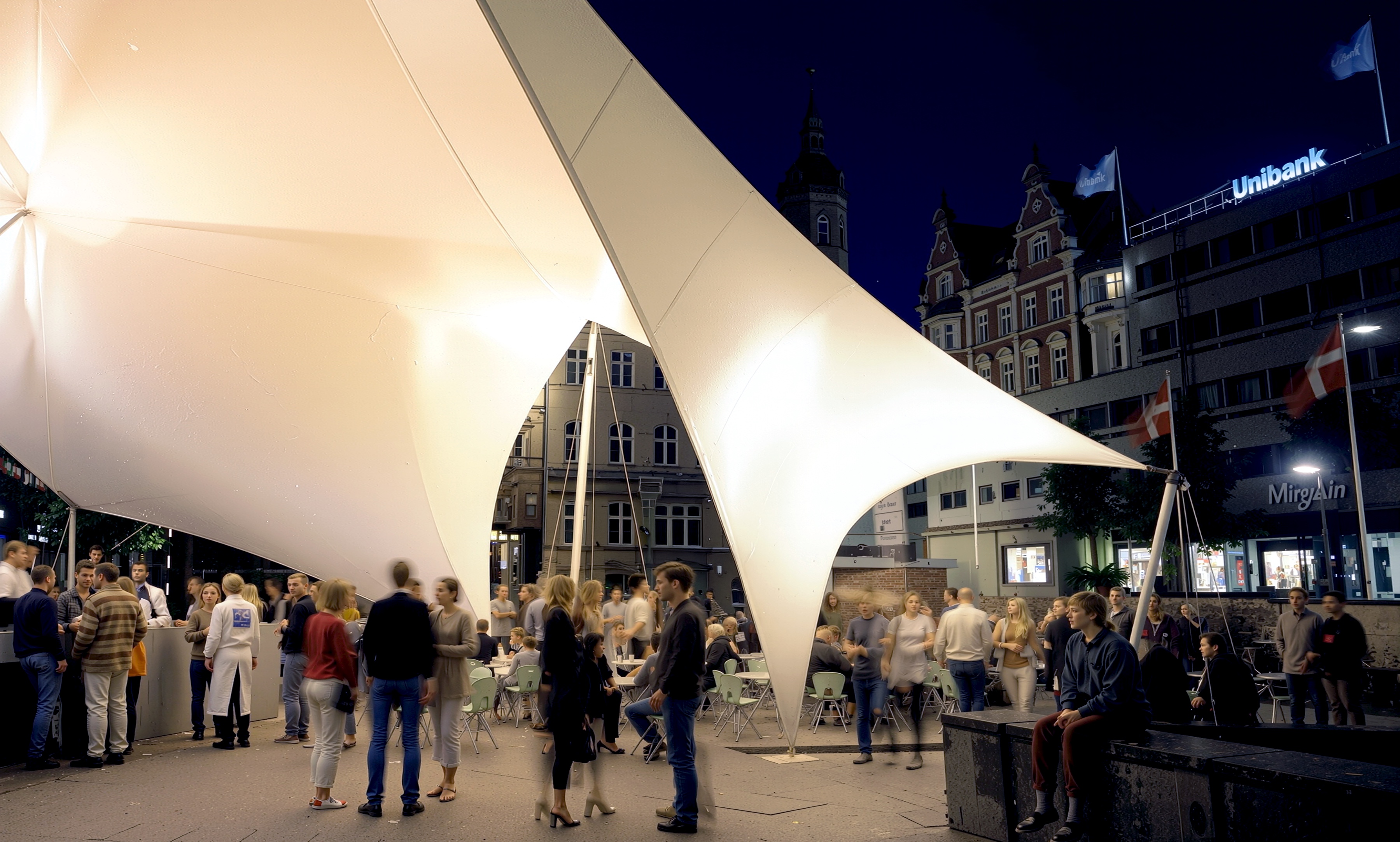 Evening view of visitors gathering beneath the illuminated tensile canopy with Aarhus city buildings in the background