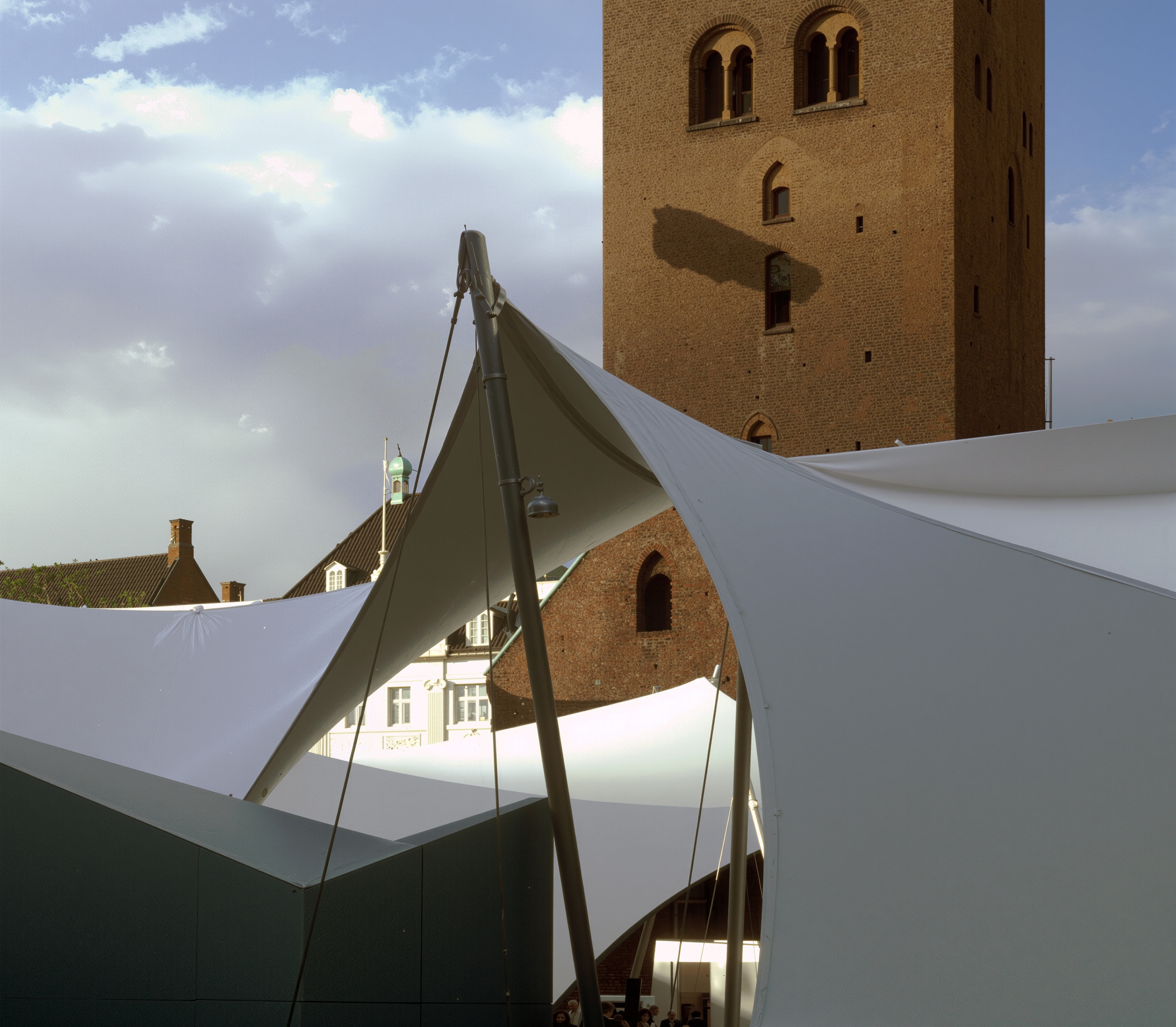 Detail of the tensile fabric canopy and steel mast structure with the historic Aarhus Cathedral tower rising behind