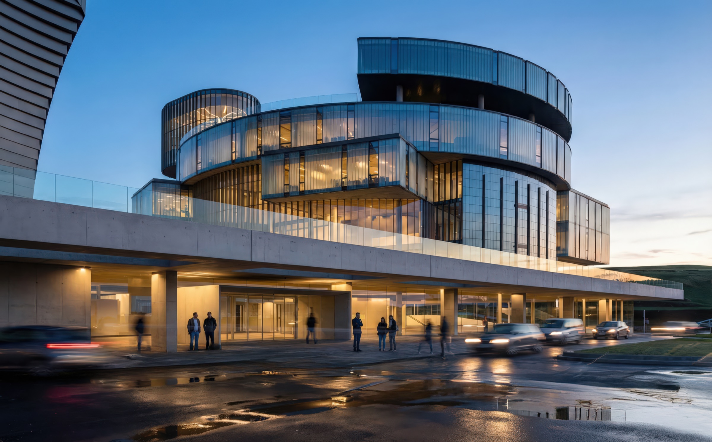 Night-time street-level view of the cultural complex showing curved glass facades wrapping the upper floors with warmly lit interiors visible through the glazing, a concrete canopy sheltering the ground-level entrance, pedestrians and vehicles on the wet street, and a dark rainy sky above
