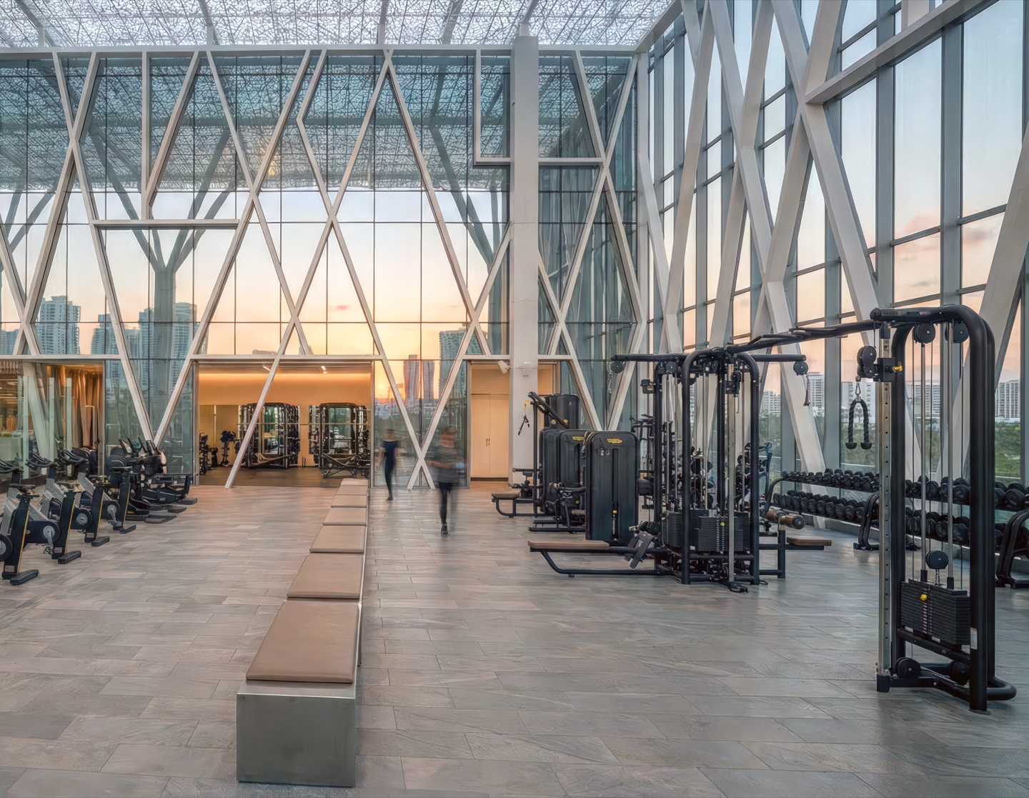 Double-height interior gym floor with weight and cardio equipment beneath a soaring diagrid glass curtain wall reflecting the Miami sunset