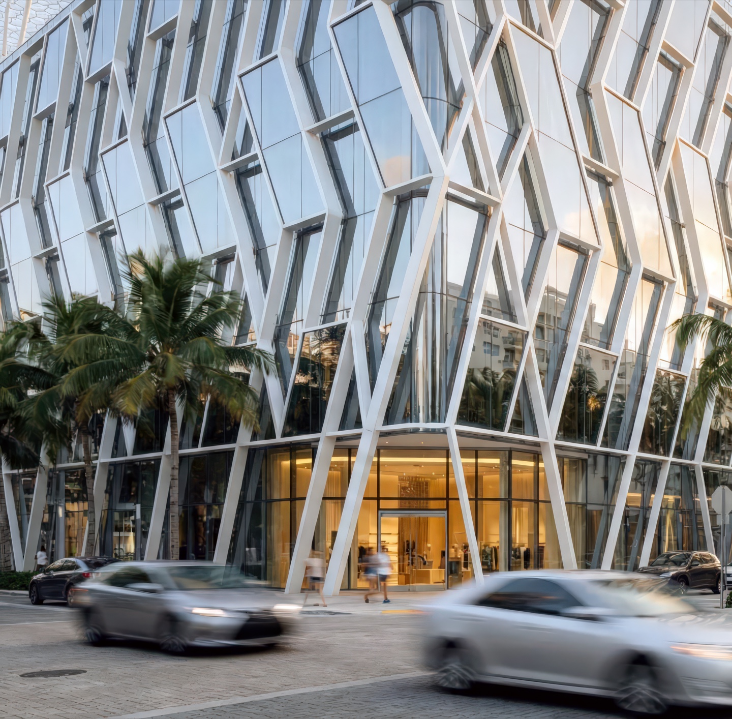 Street-level view of the diamond-patterned white structural facade with illuminated ground-floor entrance framed by palm trees and passing traffic