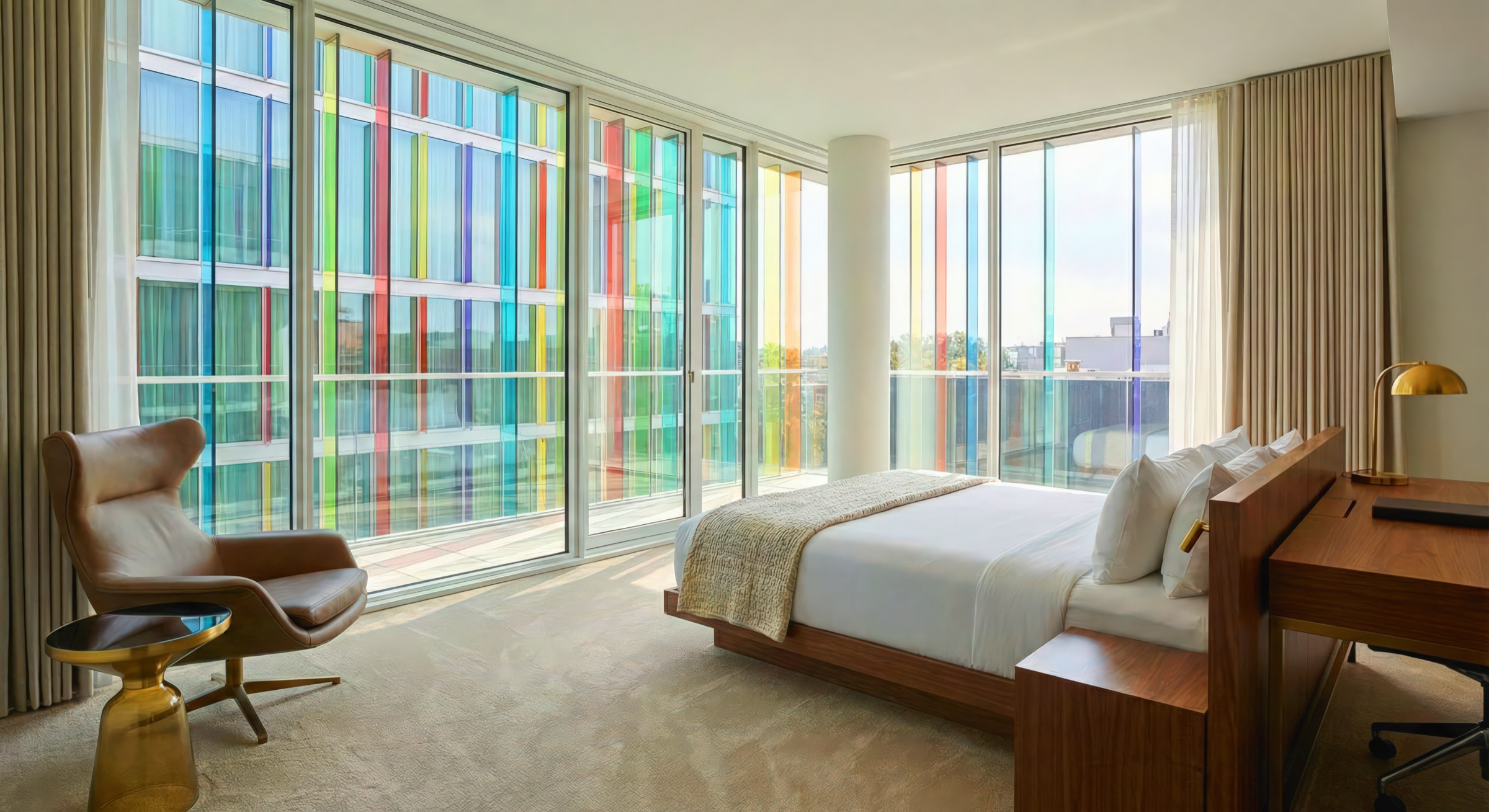 Hotel guest room with walnut platform bed, floor-to-ceiling windows revealing the colored glass facade of the adjacent wing, leather lounge chair, brass side table, and sheer curtains