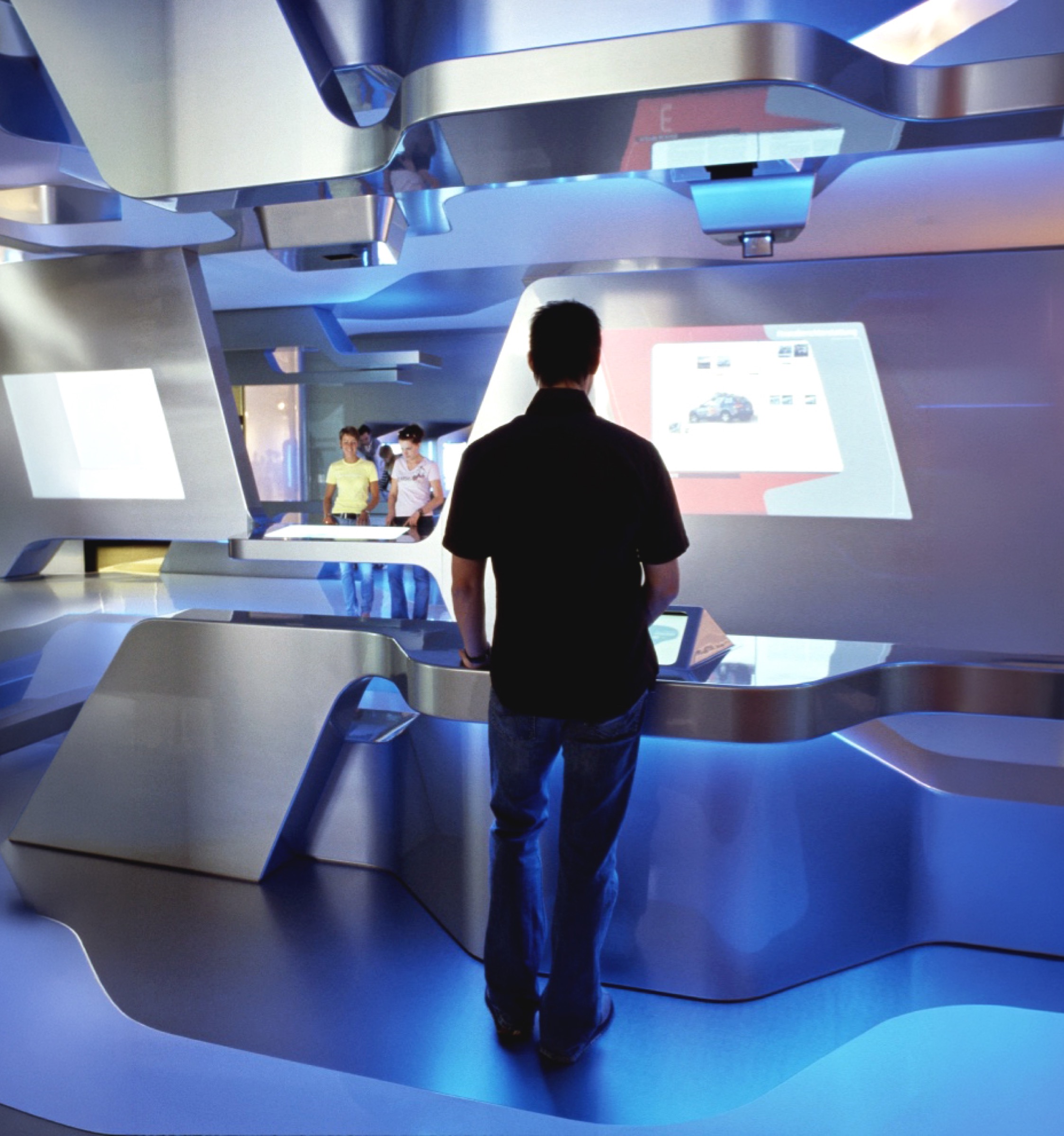 A visitor standing at a sculpted metallic console facing a projected screen showing vehicle data, angular overhead canopy with projectors, other visitors visible beyond the installation's continuous interior landscape