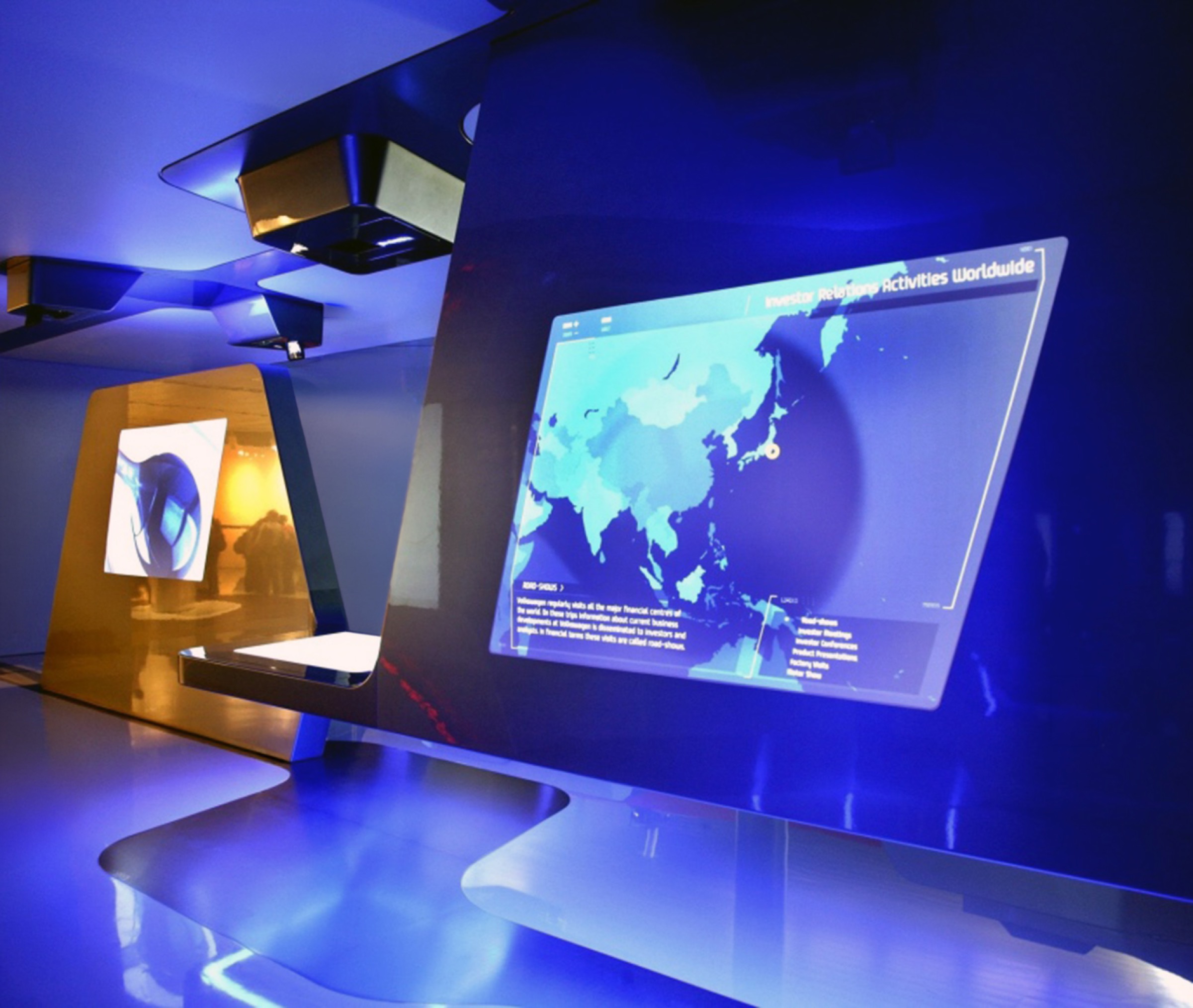 Close-up of a large projected screen displaying an interactive world map titled Investor Relations Activities Worldwide, framed by angular blue metallic canopy elements and overhead projectors, deep blue ambient lighting