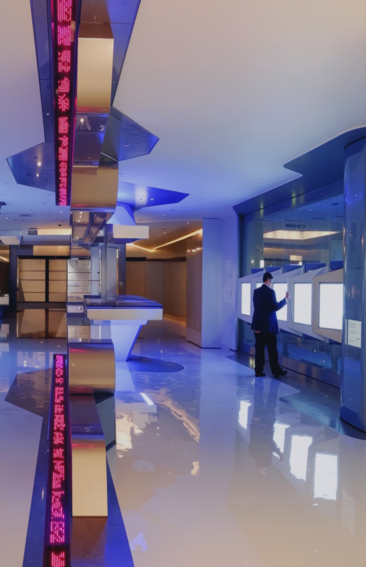 Vertical view of the main hall with an angular LED ticker column displaying red financial data, a visitor at interactive light-box terminals on the right, sculpted metallic ceiling elements, and a highly reflective floor