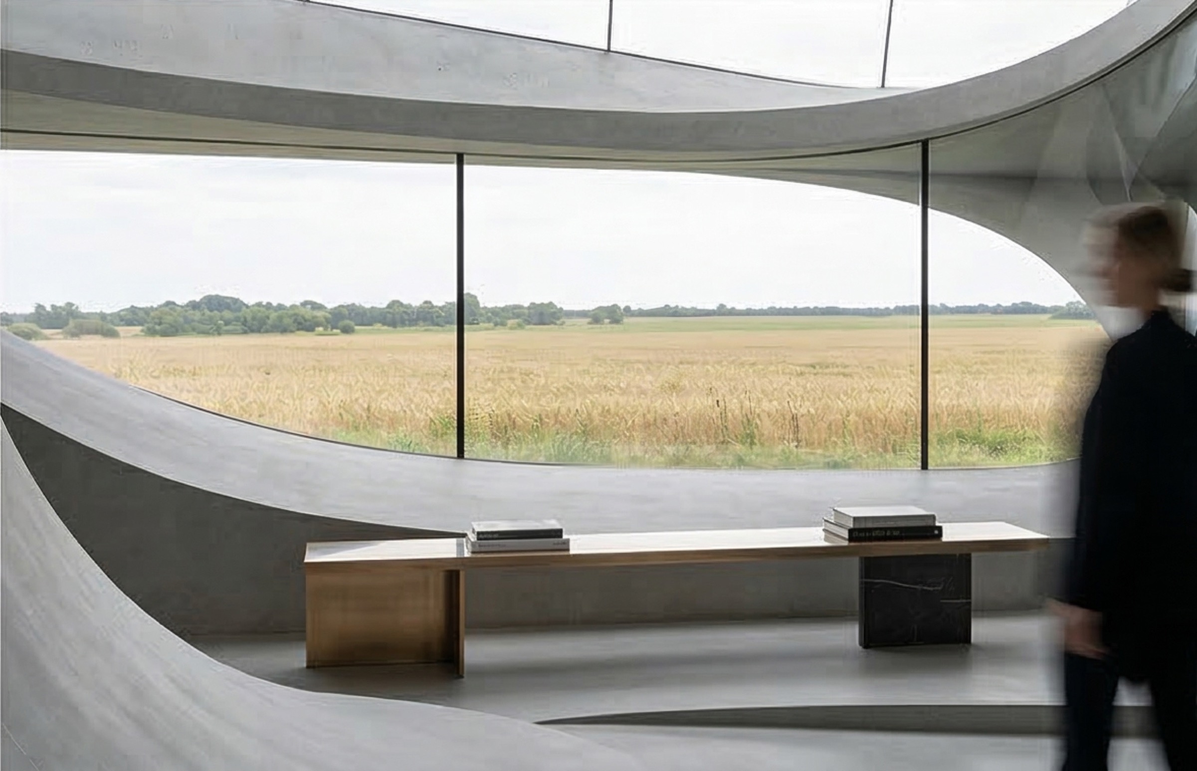 A concrete interior with a long timber-and-marble bench beneath a panoramic curved window framing golden fields and distant trees in summer