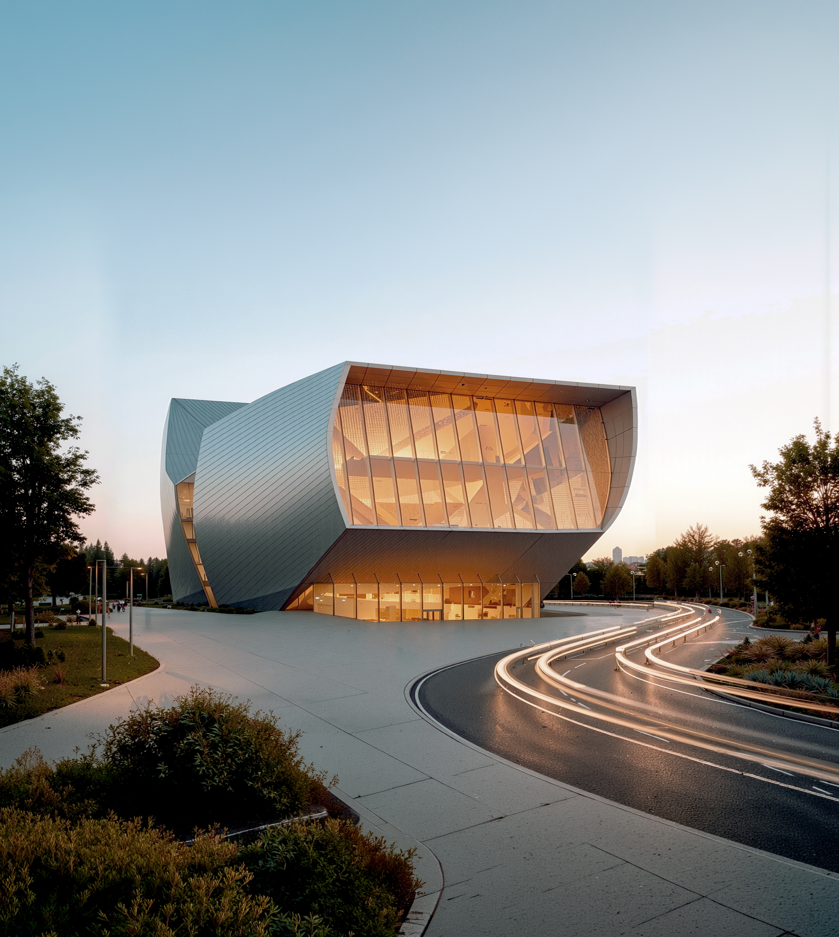 The performing arts center at dusk in summer, its faceted metallic envelope glowing warmly against a twilight sky with a sweeping approach drive